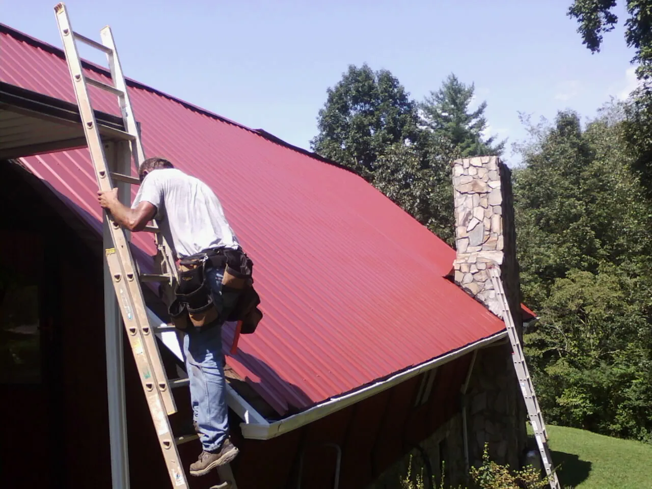 Roofer on a ladder beside a red metal roof and stone chimney — Soffit & Fascia Repair by Roofing Brothers in Thaxton